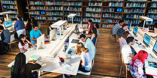 Several students working in a technology library, using computers, books, and printed materials.