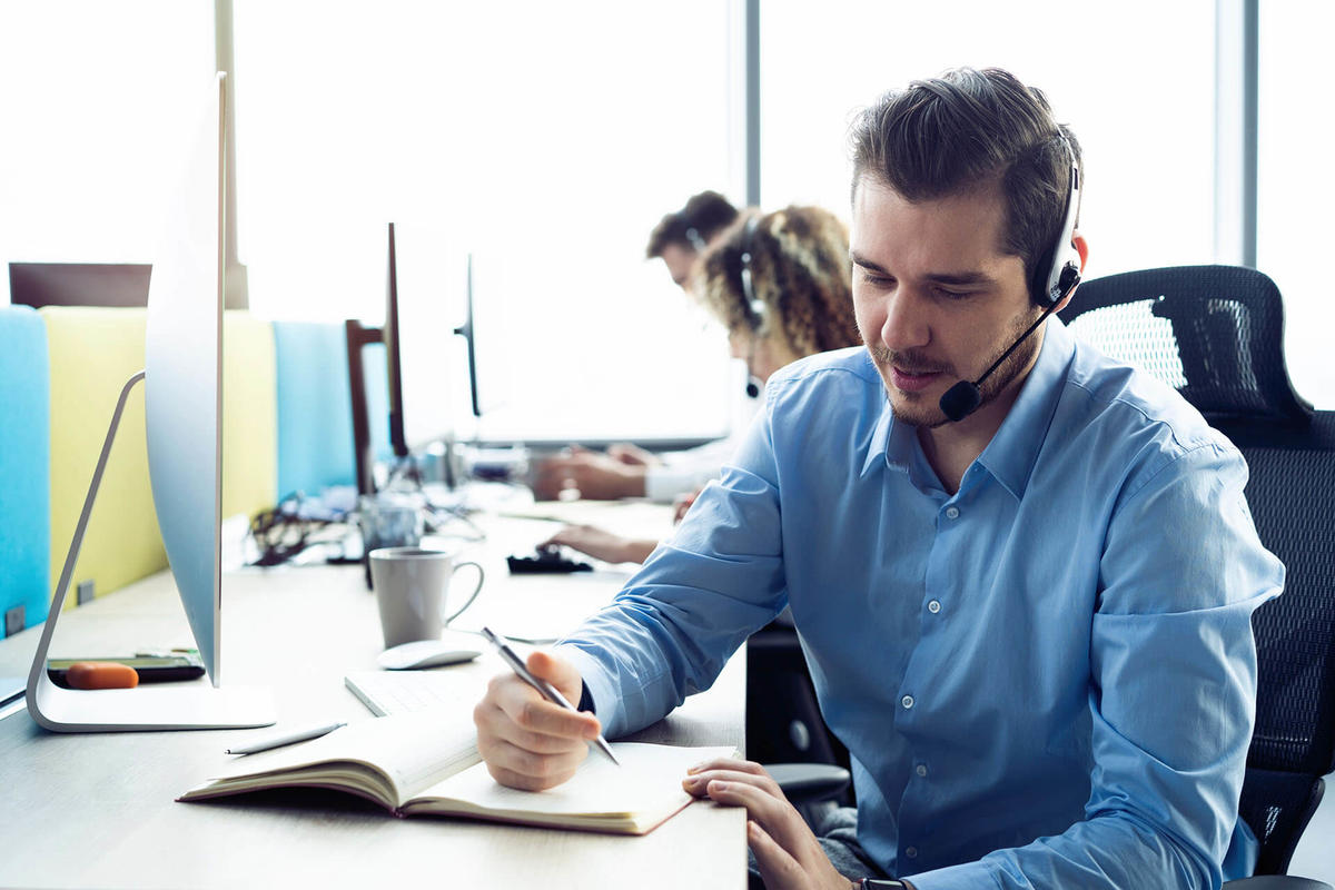 Man with headset writing in a book while working at a contact center