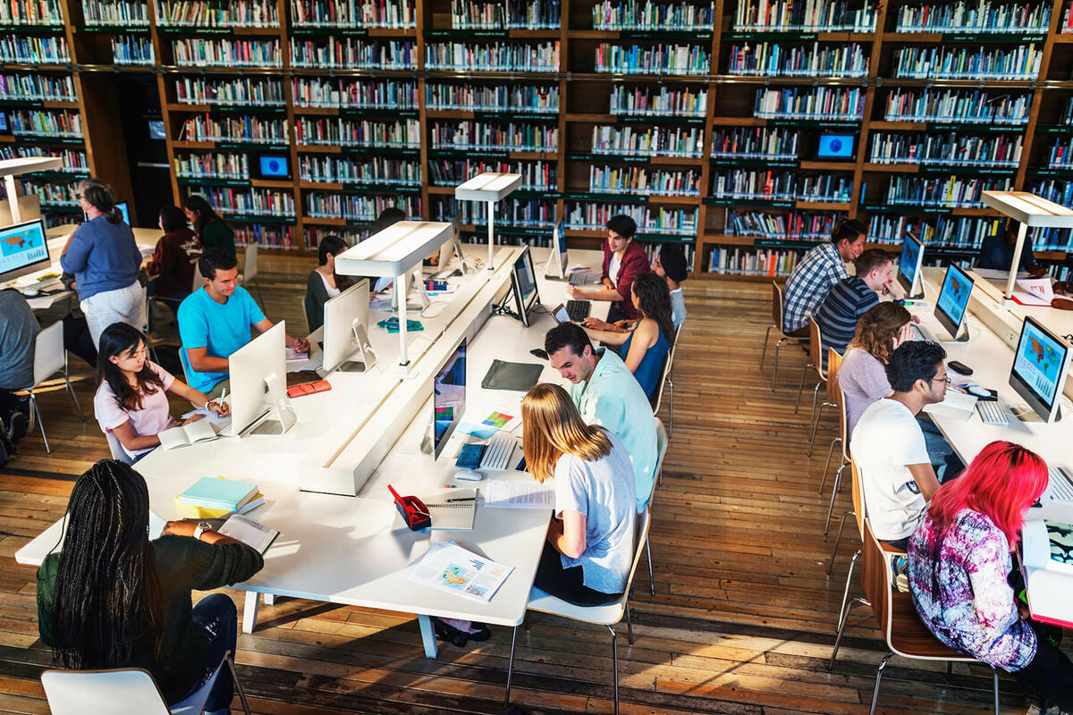 Several students working in a technology library, using computers, books, and printed materials.