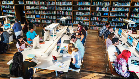 Several students working in a technology library, using computers, books, and printed materials.